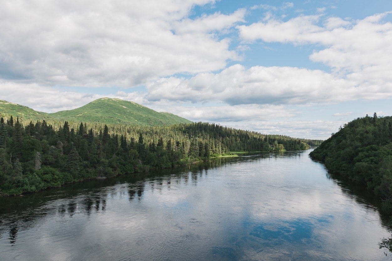 A serene river landscape bordered by lush evergreen forests and undulating hills under a dynamic sky with fluffy white clouds reflected on the water's surface.