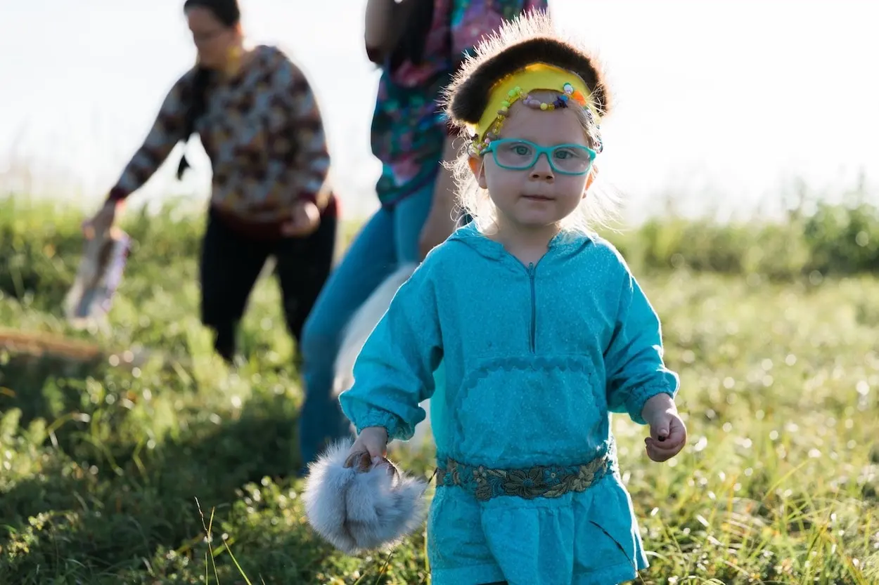 A toddler in a turquoise fuzzy onesie with glasses and a colorful headband walks in a field, holding an Alaska Native Dance Fan.
